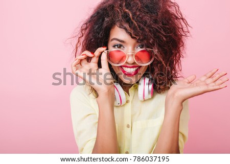 Good-looking young woman with brown skin holding pink sunglasses and posing with surprised smile. Indoor portrait of emotional african female model in elegant yellow attire.