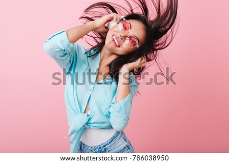 Enthusiastic brunette girl dancing with hair waving during photoshoot in studio with pink interior. Indoor photo of active hispanic woman in headphones and sunglasses having fun.