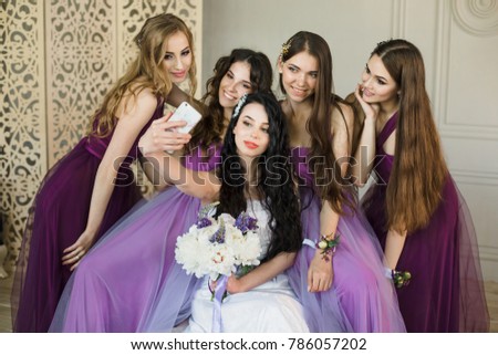 Bridesmaids taking a selfie on a smartphone and laugh during the morning of the bride at home. Beautiful girls in purple wedding dresses and bride with bouquet of peonies have fun. Preparation