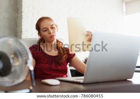 Portrait of young redhead woman working with computer laptop in office at summer during heatwave. The temperature is hot and the hair conditioner is broken. The girl sweats and feels exhausted Royalty-Free Stock Photo #786080455