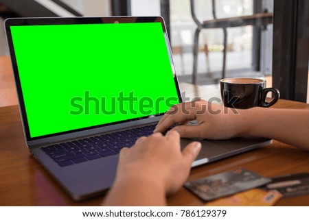 Mockup image of hands typing on laptop keyboard with blank green desktop screen and credit cards on wooden table in modern cafe