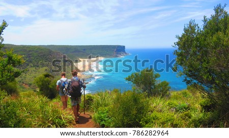 Two young men trekking through the Royal National Park, near Sydney Australia with sticks and backpacks on with a lovely view of Gerai Beach ahead on top of a hill track                          Royalty-Free Stock Photo #786282964