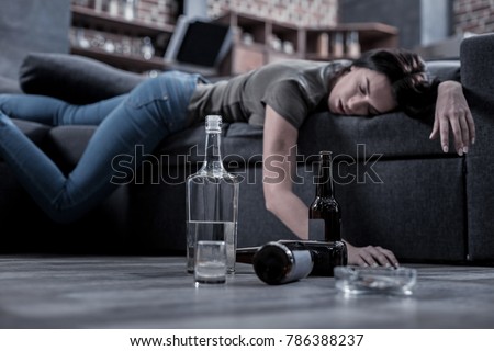 Unfinished drink. Selective focus of half empty bottle of vodka standing on the floor with a drunken young woman sleeping in the background Royalty-Free Stock Photo #786388237