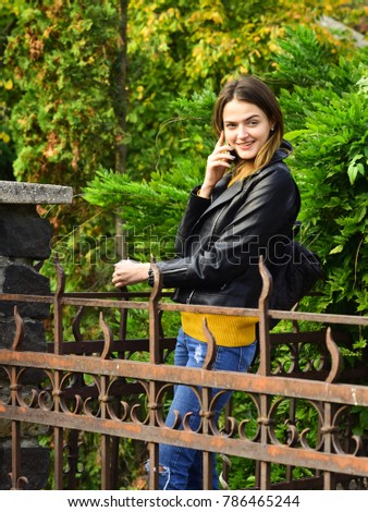 Woman with tricky smiling face on green trees background. Summer or autumn season and beauty concept. Girl with pretty hair and backpack enjoys fall time. Tourist posing near metal figure fence