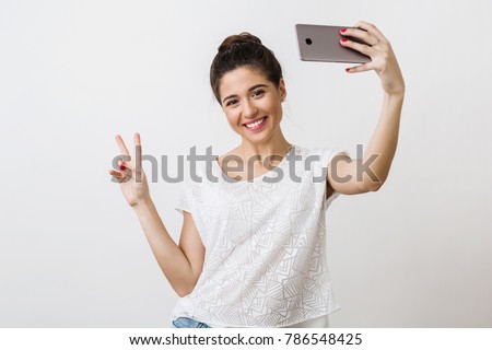 young attractive natural smiling woman in white blouse holding smart phone, showing peace sign, positive emotion, making selfie photo, looking into camera, isolated