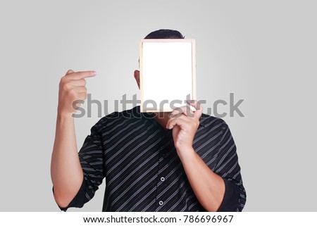 Portrait of a man wearing black shirt, holding and hiding covering his face with small empty whiteboard, pointing on copy space mask