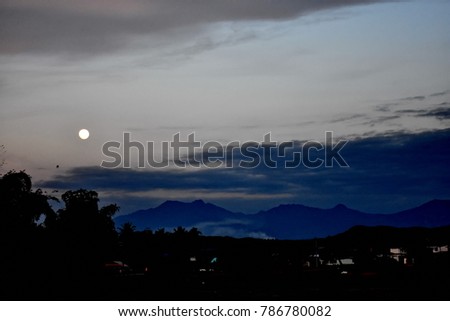 Silhouette picture of village in the countryside at Phrae province of Thailand with mountain, clouds, dim light sky and full moon on January 2018 with a little bird.