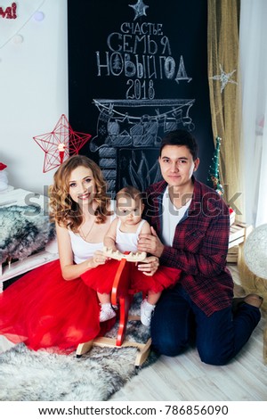 Happy cute little baby girl on rocking dear at Christmas tree with a toy, dressed in red dress New Year room at home. Child good mood. Lifestyle, family and holiday concept. With parents