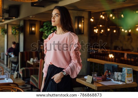 Long-haired brunette girl in cafe. Standing. Pink turtleneck and black skirt. Lovely interior of the city cafe. confident view of the future, motivation. Place for text.
