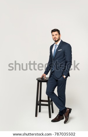 handsome serious young businessman leaning at stool and looking at camera isolated on grey