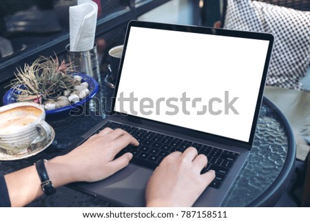Mockup image of hands using and typing on laptop with blank white desktop screen and coffee cup on vintage wooden table in cafe