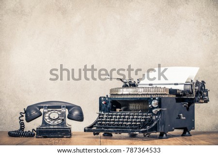 Retro old black telephone and aged typewriter with paper blank on wooden desk front concrete wall background. Vintage style filtered photo