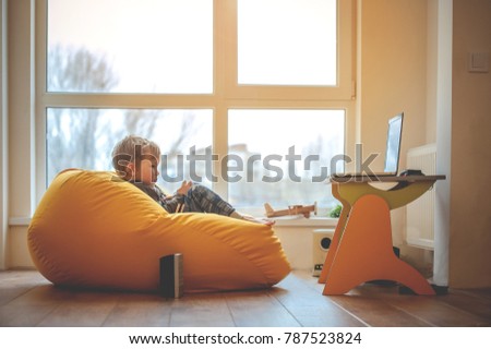 Child and laptop. Little boy on yellow bean bag at home interior watching cartoons on laptop. Addiction 