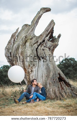 The young romantic couple in love sitting in embrace outdoor and relying on the trunk of tree and boyfriend holding the white balloon in hand
