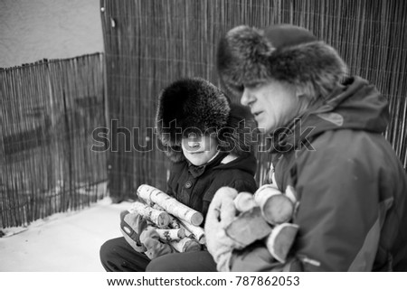 Child and grandfather in fur hats with birch woods in hands sitting outside near house. Cold russian winter. Black and white photo