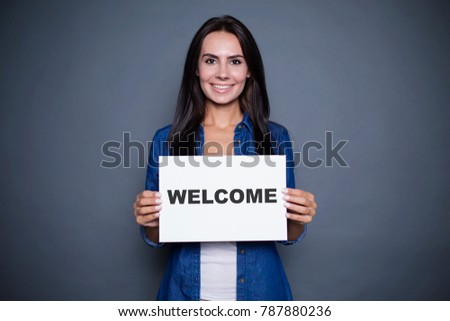 Welcome at new home. Beautiful modern smiling woman in a jeans shirt is holding a sign with the inscription "welcome" on a gray background isolated.