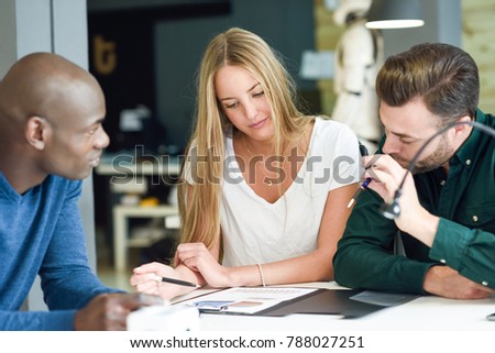 Three young people studying with graphics on white table. Beautiful blonde girl, african and caucasian men working together wearing casual clothes. Multi-ethnic group.