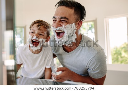 Man and little boy with shaving foam on their faces looking into the bathroom mirror and laughing. Father and son having fun while shaving in bathroom. Royalty-Free Stock Photo #788255794