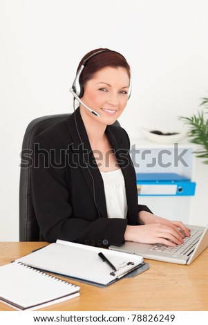 Young gorgeous red-haired woman in suit typing on her laptop and using headphones while sitting in an office