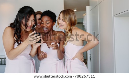 Smiling bridesmaids looking at the photographs of bride on mobile phone in a hotel room on wedding day. Beautiful bride and bridesmaid on the wedding day