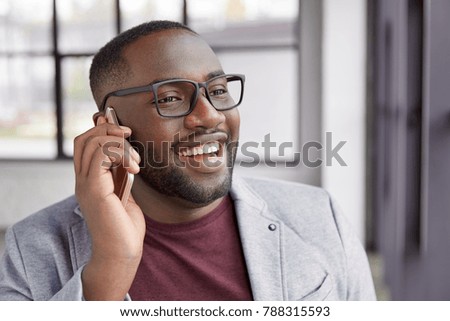 Photo of cheerful black male with shing smile, has white perfect teeth, calls secretary, dressed formally, stands in spacious cabinet. Handsome African American male financier has phone conversation
