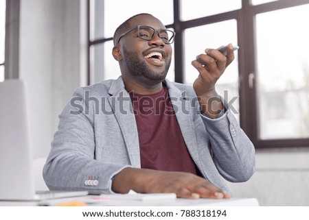 Cheerful dark skinned male makes voice call, happy to hear business partner, share financial news and successful signed contract, sits over working interiror against big window, smiles joyfully