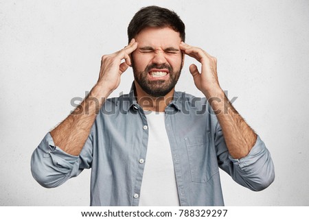 Frustrated handsome young male clenches teeth with pain, has headache, keeps hands on temples, feels terrible after loud noisy party, needs calm atmosphere, isolated over white studio background