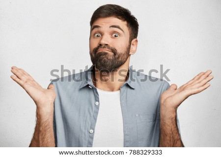 Maybe yes or no. Doubtful bearded fashionable guy shrugs shoulders in bewilderment, tries to make decision, puzzled what he wants, isolated over white concrete wall. Hesitation and uncertainty Royalty-Free Stock Photo #788329333