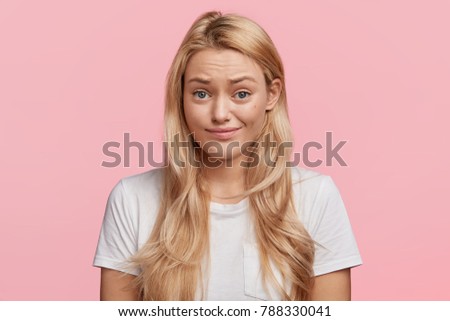 Adorable blonde young woman without make up, looks with displeased expression, raises eyebrows, can`t understand something, isolated over pink background. People and facial expressions concept