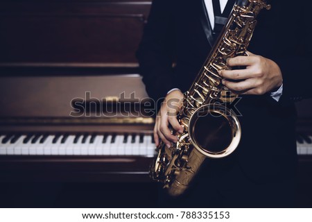 close up of Young Saxophone Player hands  playing alto sax musical instrument over the piano  background,  closeup with copy space, vintage tone,  can be used for music background Royalty-Free Stock Photo #788335153