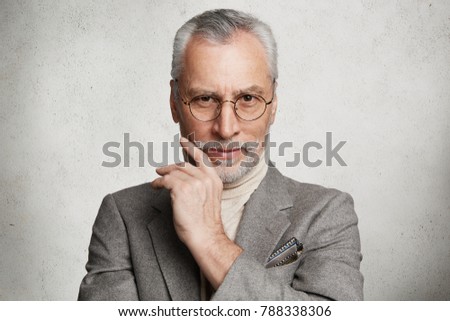 Pleased handsome wrinkled mature man wears spectacles and formal suit, looks directly into camera with confident expression, being glad to be promoted, isolated over white concrete background Royalty-Free Stock Photo #788338306
