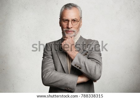 Serious senior male magnat dressed in formal suit, round spectacles, keeps hands crossed, listens attentively financial report, isolated over white concrete background. Pensioner poses indoor Royalty-Free Stock Photo #788338315
