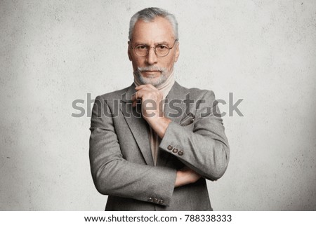 Indoor shot of prosperous experienced elderly wrinkled businessman dressed in grey formal suit, keeps hand under chin, comes for business meeting, isolated over white studio concrete background. Royalty-Free Stock Photo #788338333