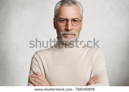 Photo of intelligent serious bearded mature man keeps hands crossed, wears light poloneck sweater and glasses, spends free time in circle of friends, isolated over white concrete background. Royalty-Free Stock Photo #788340898