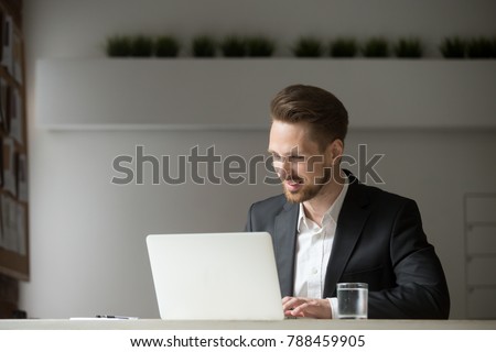 Smiling attractive businessman working online on computer sitting in suit at office desk, young executive manager looking at laptop screen at workplace, happy man using app for business on pc concept