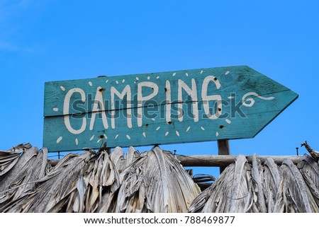 Blue handmade handwritten painted vintage camping sign made out of weathered old wood showing in to the right side direction on a palapa palm tree roof with blue sky background
