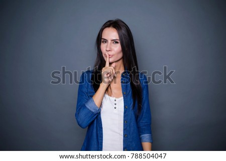 Can you be quiet? A beautiful concentrated woman in a denim shirt puts her index finger to her lips on a gray background isolated.