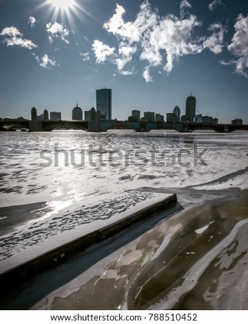 Pier on frozen river with buildings in background made of concrete and steel, encased in glass reach towards cloud filled  sky.