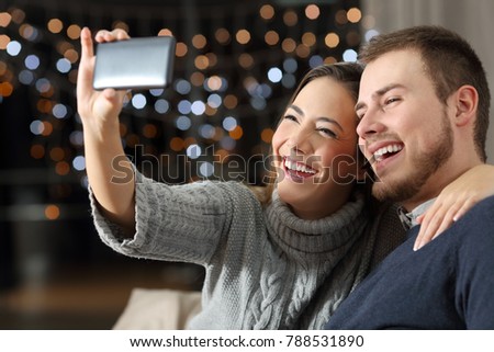 Happy couple taking selfies with a smart phone sitting on a couch in a winter night at home