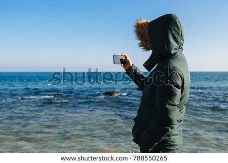 Young man taking a picture of the seascape on smartphone, outdoors. Back view.