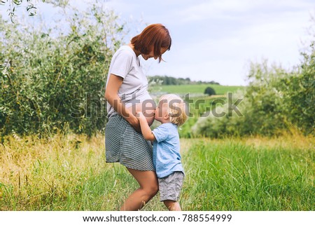 Child boy hugging and kissing belly of pregnant her mother against green nature background. Pregnant woman and her toddler son outdoors. Pregnancy, family, parenthood concepts.