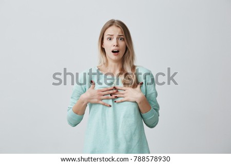 Surprised terrified blonde female gestures with uncertainty, stares at camera, puzzled as doesn`t know anwser on question, isolated against gray background. People, body language, emotions concept
