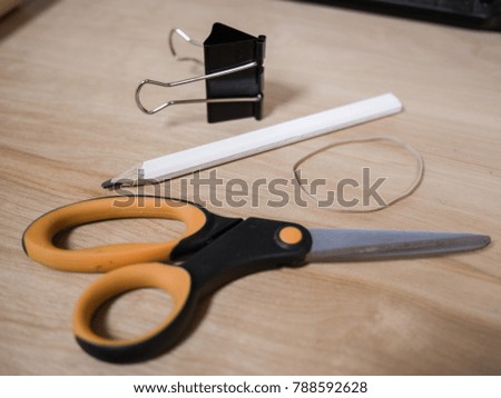 Close up photograph of office supplies including a scissors, rubber band, a white carpenter pencil, and a black metal binder clip on top of a light colored wood grain desk.