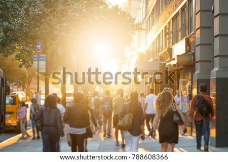Man stands in the middle of a busy sidewalk looking at his cell phone while crowds of people walk around on 14th Street in Manhattan, New York City with the glow of sunlight in the background. Royalty-Free Stock Photo #788608366