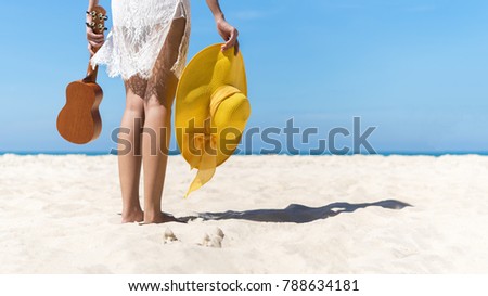 Close up of young lady holding ukulele and yellow beach hat on the beach with blue sky in sunny day with free space for text. Travel and summer background concept.