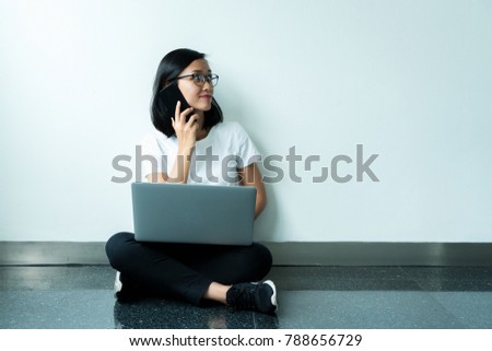 smiling beauty girl student sitting on floor with white wall background and talking mobile phone looking at air daydreaming when she using laptop computer study.