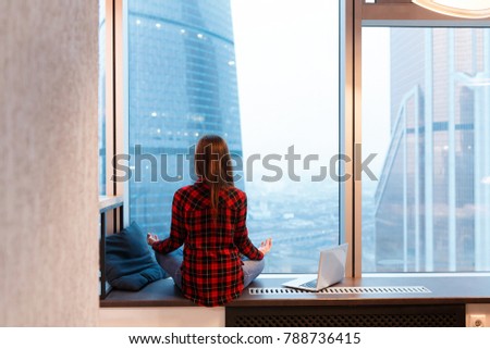 Young unidentified woman freelancer designer in casual clothes admiring the view and meditating during break in the background of window overlooking skyscrapers