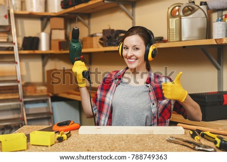Beautiful caucasian young brown-hair woman in plaid shirt, noise insulated headphones working in carpentry workshop at table place, drilling with power drill holes in piece of wood, making furniture