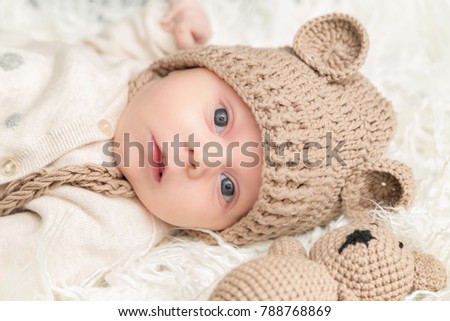 Newborn baby with knitted cap and bear laying white fur background. Baby portrait.