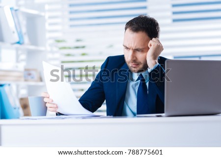 Reading. Clever skilled experienced manager sitting at the table with his head leaning on his hand and feeling bored while reading the documents